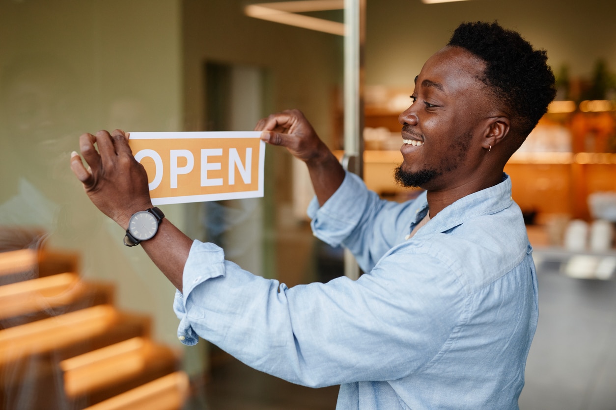 Joyful young African American man opening his small business cafe in morning sticking sign on glass doo